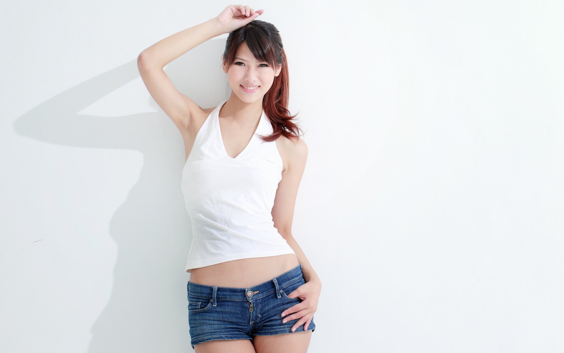 A woman model in a white top and denim shorts poses confidently against a plain white wall, featured in a high-definition desktop wallpaper background.