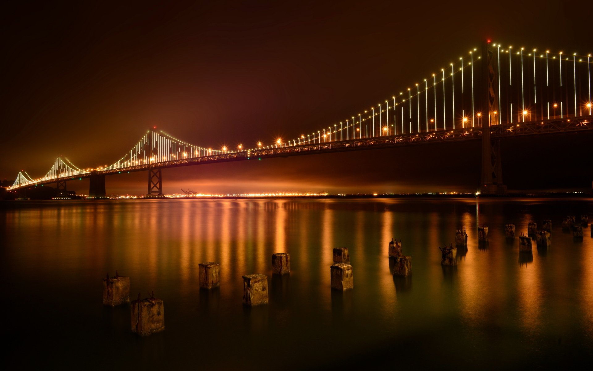 Night view of the illuminated San Francisco Bay Bridge reflecting on calm waters, captured in HD as a striking desktop wallpaper background.