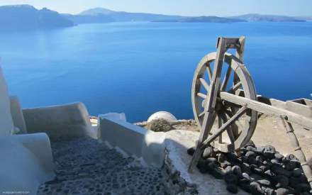 A stunning panorama of the blue sea in Santorini, Greece, featuring man-made structures alongside cobblestone pathways and a rustic wooden wheel by the water's edge.