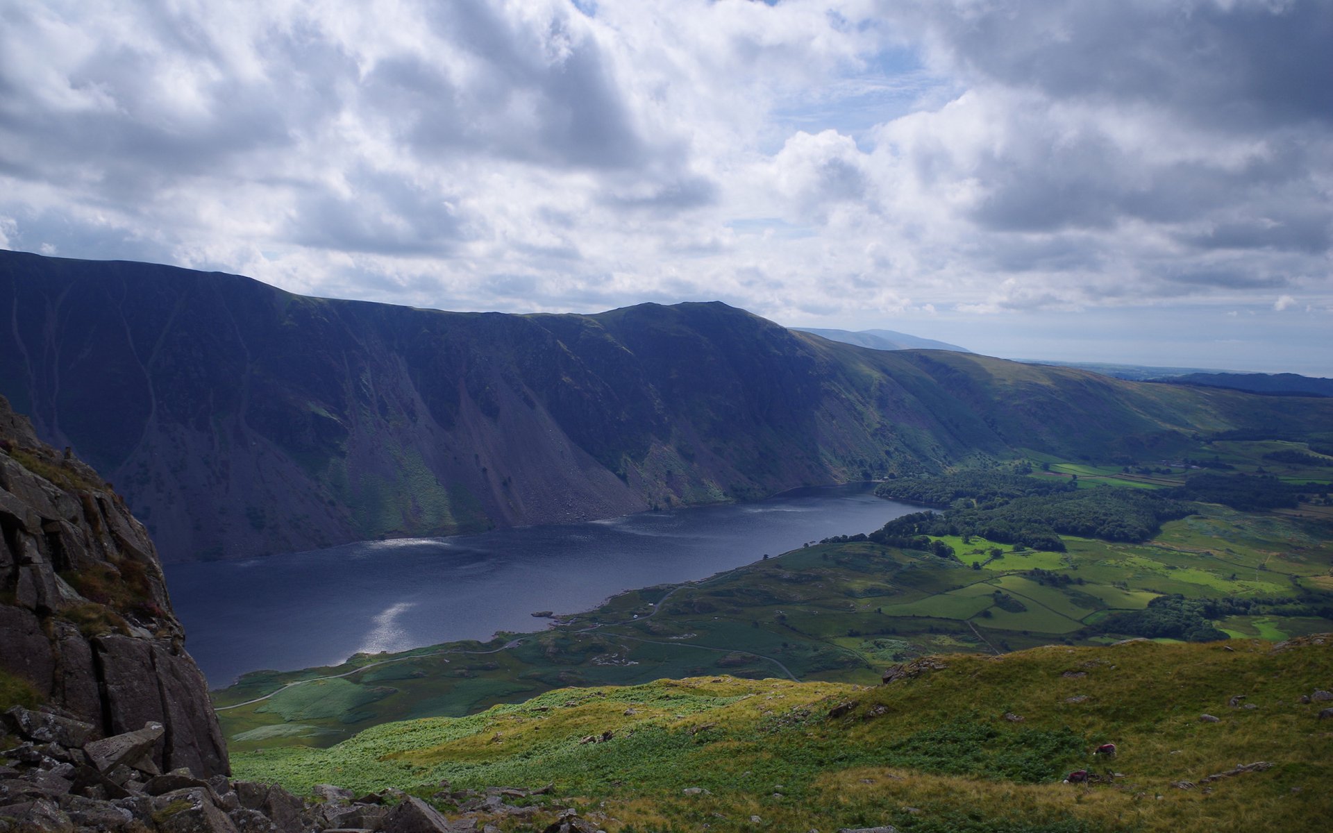 2K Quad HD PC desktop background: panoramic mountain landscape with a deep glacial lake cradled by steep cliffs and rolling green valleys under a dramatic cloud-filled sky.