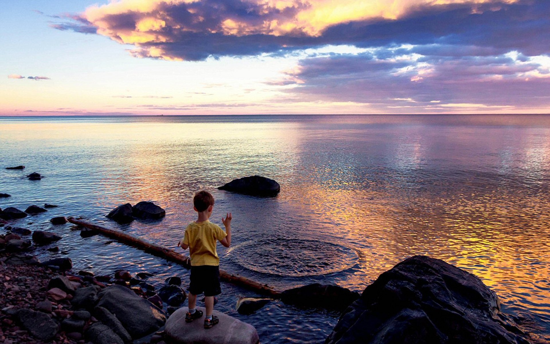 Photography: child in yellow shirt standing on rocks at sunset, making ripples in calm, colorful sea — vibrant sky reflections; HD PC desktop wallpaper and background.