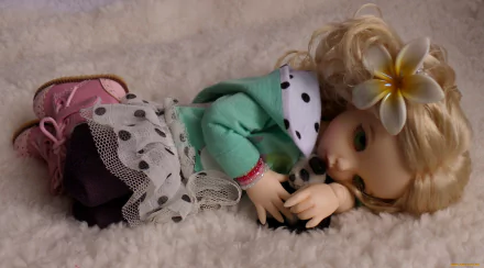 HD desktop wallpaper featuring a detailed man-made doll lying on soft fabric, dressed in layered clothing with a flower in its blonde hair.