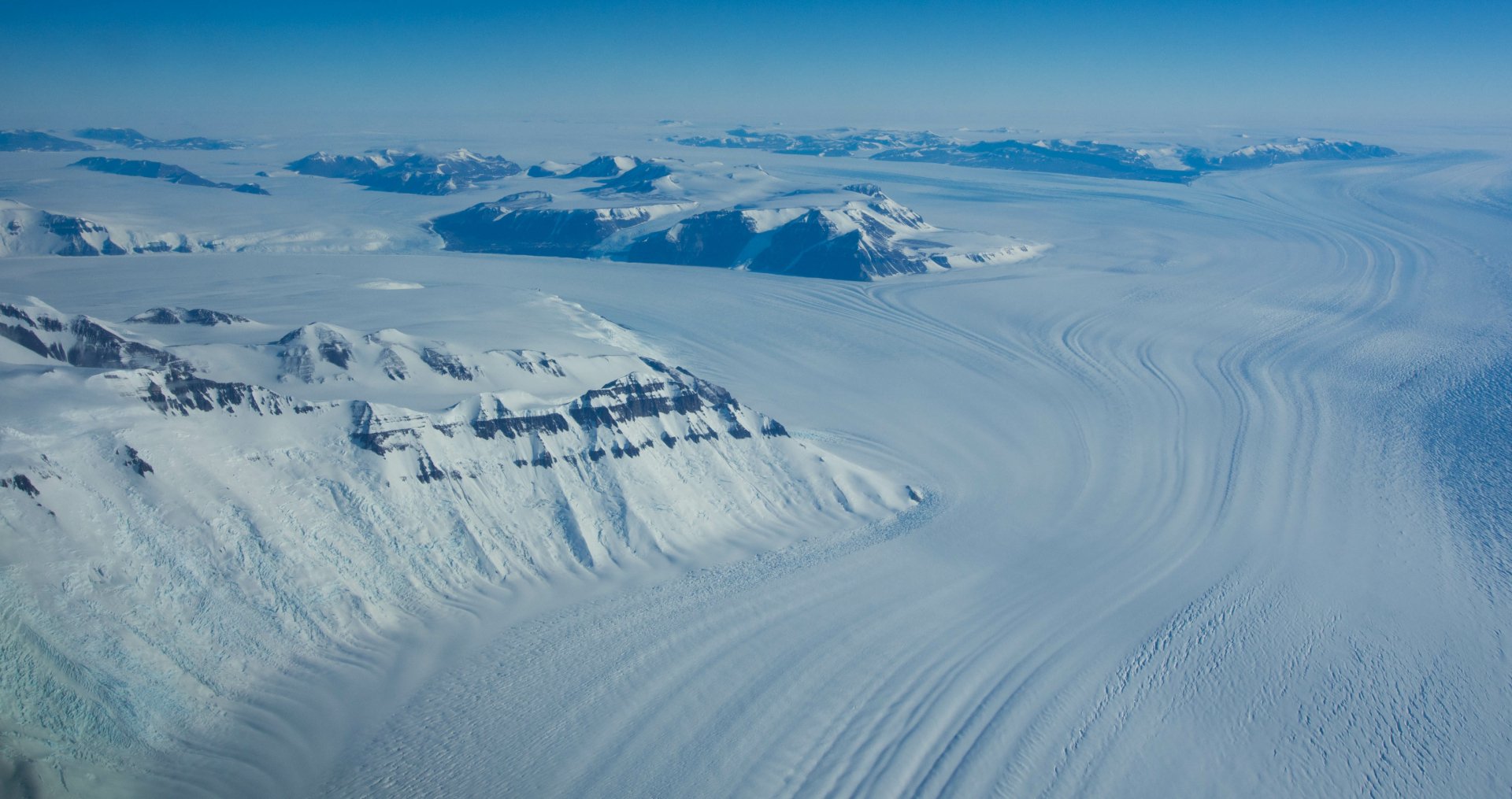Aerial view of McMurdo Station man-made outpost beside vast Antarctic ice plains and ridged glaciers, snow-clad mountains on the horizon — 4K Ultra HD PC desktop wallpaper.