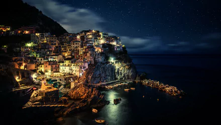 HD wallpaper of Manarola, Cinque Terre, Italy, at night showcasing vibrant lights of the town against the backdrop of the ocean and starlit sky.