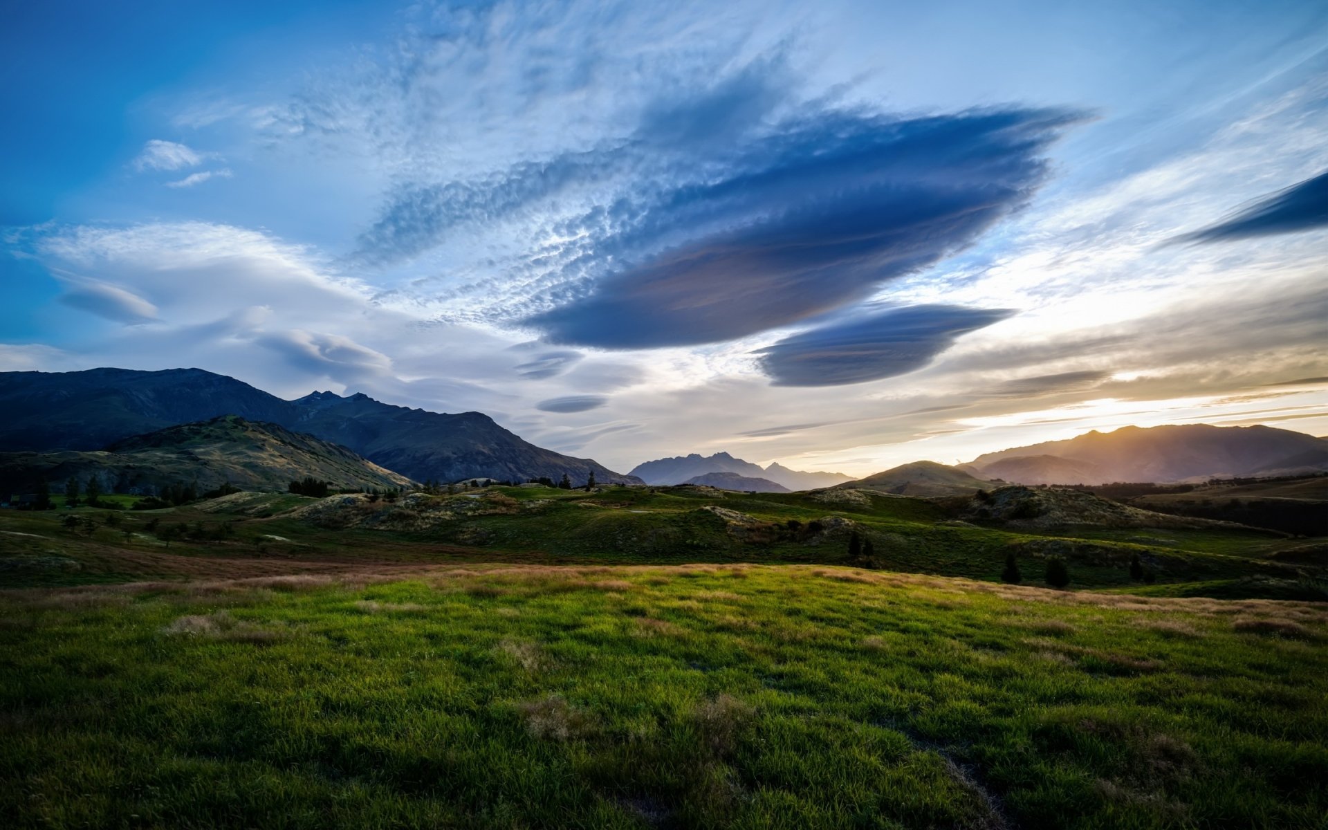 HD PC desktop wallpaper showcasing a vibrant green landscape under a dramatic sky with layered clouds and distant mountains at sunset.