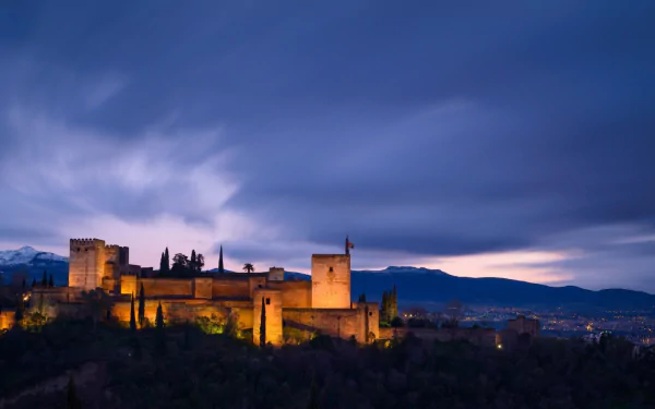 Night view of Granada's Albayzín district with illuminated historic buildings set against mountain and cityscape under a moody sky, captured in HD.