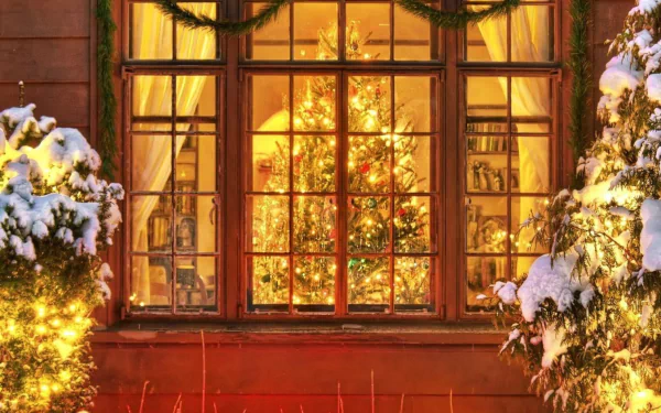 HD wallpaper of a warmly lit window with a Christmas tree and festive lights, framed by snow-covered branches.
