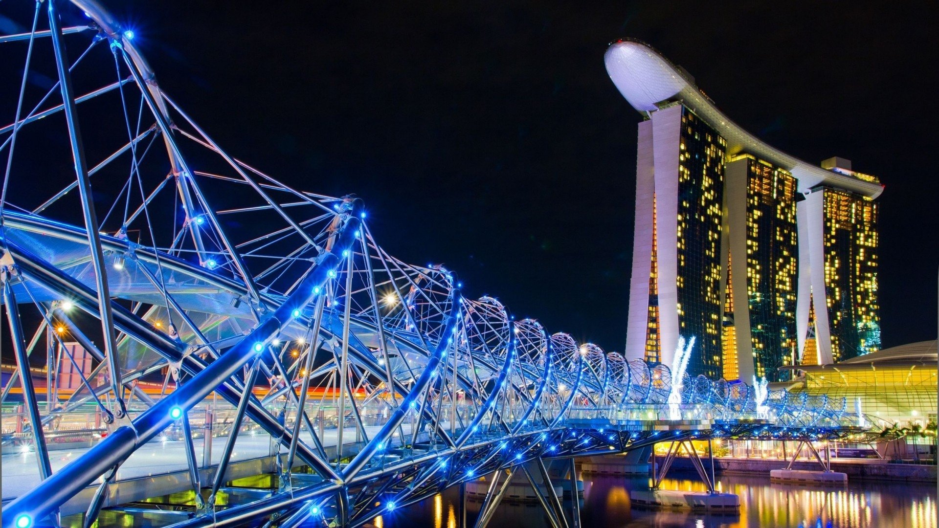 Night view of Singapore's Helix Bridge illuminated in blue, with the iconic Marina Bay Sands hotel glowing in the background.