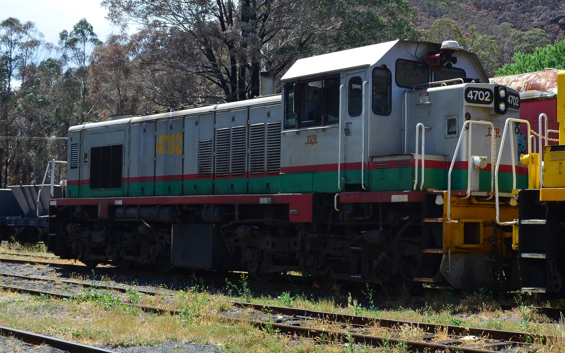 Lithgow diesel locomotive train vehicle on sunlit tracks, green-gray body with red stripe and yellow accents, trees behind — HD PC desktop wallpaper and background.