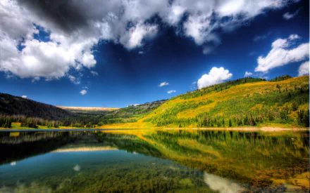 A serene lake surrounded by lush greenery and majestic hills under a vibrant blue sky with scattered clouds, capturing the tranquility of nature in this HD desktop wallpaper.
