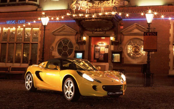 HD desktop wallpaper featuring a yellow Lotus Elise sports car parked in front of a warmly lit music café at night.