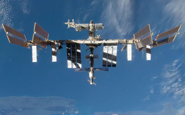 High-definition image of the International Space Station orbiting Earth, showcasing its solar panels and intricate structure against a backdrop of clouds and blue sky.