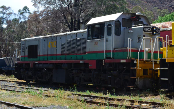Lithgow diesel locomotive train vehicle on sunlit tracks, green-gray body with red stripe and yellow accents, trees behind — HD PC desktop wallpaper and background.