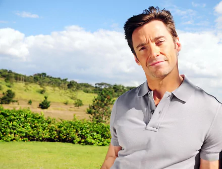 Hugh Jackman poses outdoors in a grey polo shirt against a scenic background of green hills and a bright blue sky, making this an eye-catching HD desktop wallpaper.