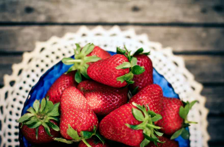 HD desktop wallpaper featuring a close-up of fresh strawberries in a blue bowl on a lace doily and rustic wooden surface.