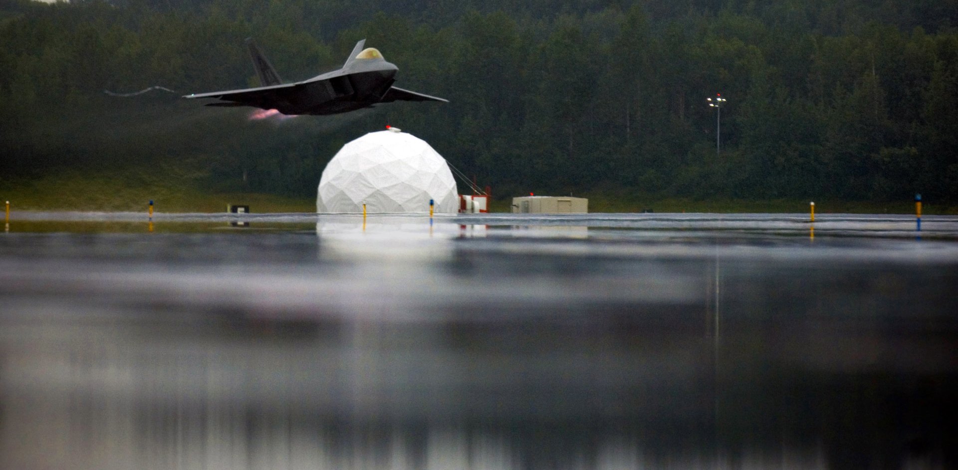 A Lockheed Martin F-22 Raptor soars above reflective water, showcasing military aviation against a backdrop of a geodesic dome and a lush green landscape.