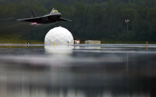 A Lockheed Martin F-22 Raptor soars above reflective water, showcasing military aviation against a backdrop of a geodesic dome and a lush green landscape.