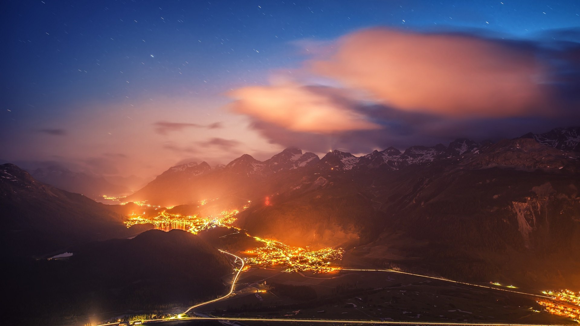 A breathtaking night landscape showcasing a valley illuminated by city lights, framed by majestic mountains beneath a dreamy sky. This 4K Ultra HD wallpaper captures serene beauty.