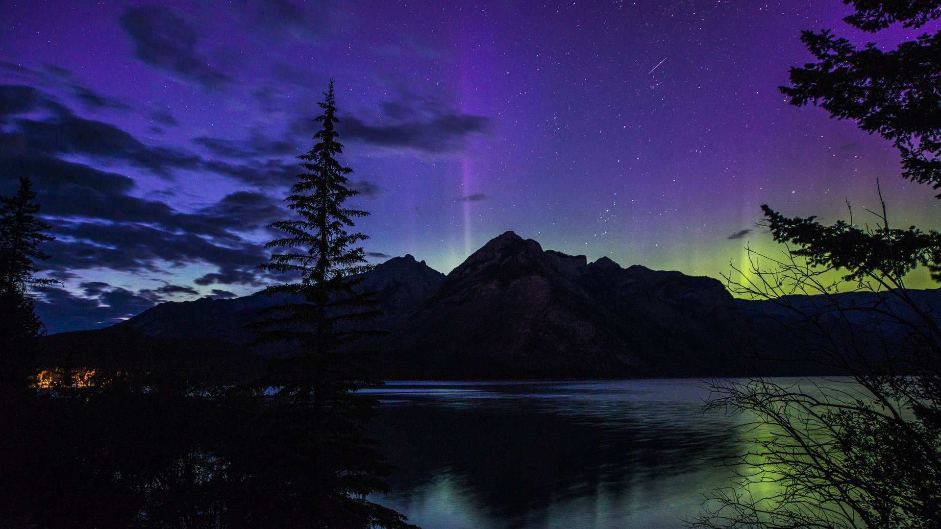 A serene landscape featuring the vibrant aurora borealis illuminating the night sky above a tranquil lake, framed by silhouetted trees and majestic mountains. A stunning nature-themed wallpaper.