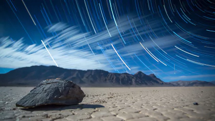 A serene view of Death Valley under a starry night sky, featuring a foreground rock and streaks of light from stars moving over the dramatic landscape, in stunning 4K Ultra HD quality.