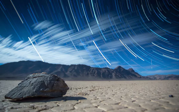 A serene view of Death Valley under a starry night sky, featuring a foreground rock and streaks of light from stars moving over the dramatic landscape, in stunning 4K Ultra HD quality.