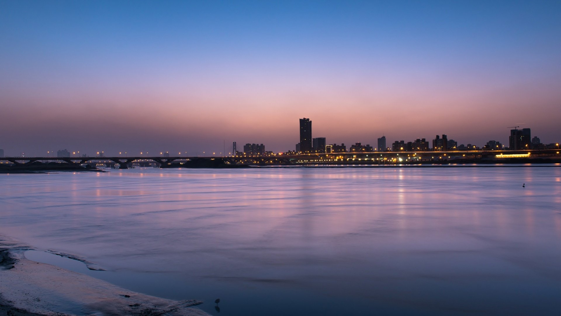 4K Ultra HD cityscape wallpaper showcasing a calm waterfront at dusk with illuminated buildings and a clear gradient sky above.