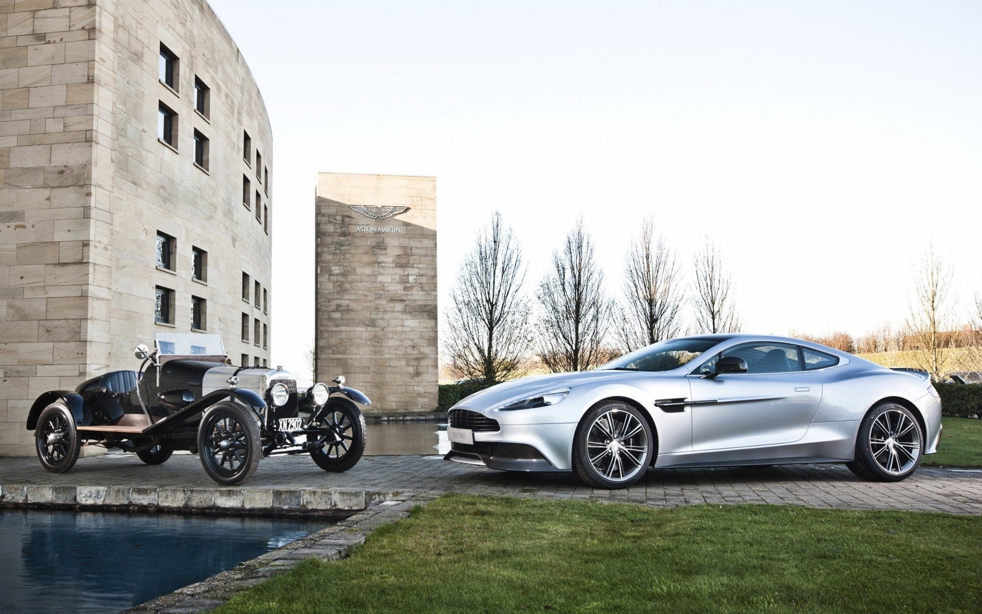 HD PC desktop wallpaper: silver Aston Martin V12 Vantage parked beside a vintage car by a modern stone building, reflecting pool, and leafless trees.