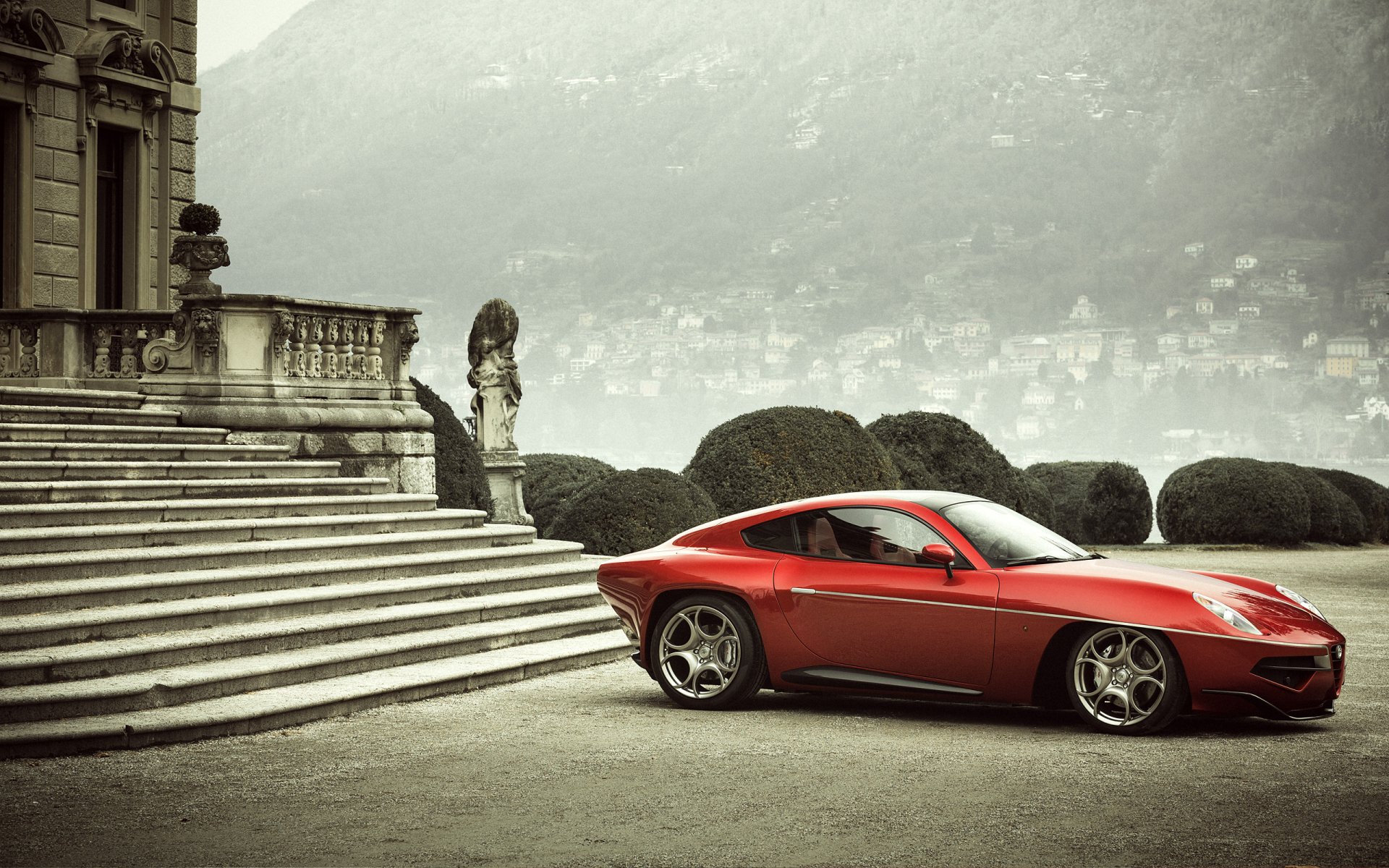 Red Alfa Romeo Disco Volante parked by grand stone steps and trimmed hedges with a misty mountain-lake backdrop — 2K Quad HD PC desktop wallpaper featuring the Alfa Romeo vehicle.