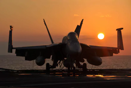 A Boeing F/A-18E/F Super Hornet warplane jet fighter is silhouetted on a carrier deck against a glowing orange sunset, captured in 4K Ultra HD quality.