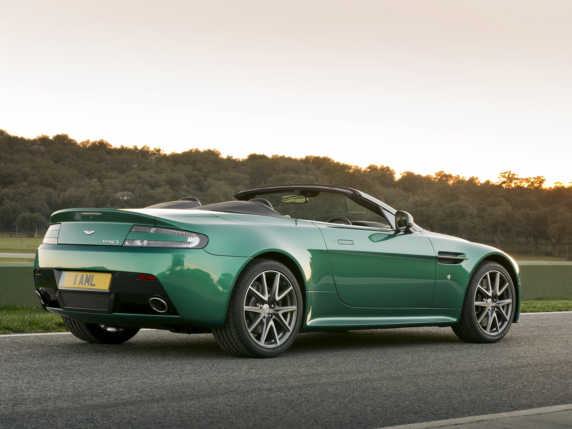 HD PC desktop wallpaper of a green Aston Martin V8 Vantage convertible parked on a roadside at sunset, shown from a rear three-quarter view.