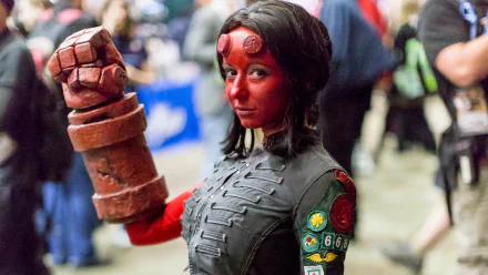 A woman in detailed Hellboy cosplay, with red paint and a large mechanical arm, stands confidently amid a lively convention backdrop. HD desktop wallpaper and background.