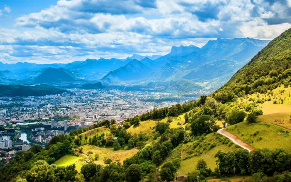 HD desktop wallpaper showing a man-made cityscape of Lyon nestled between green hills and mountains under a partly cloudy sky.