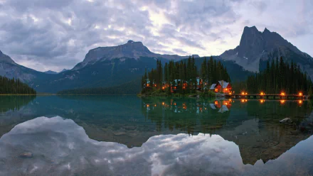 A serene landscape showcases a lake reflecting surrounding mountains and a cozy lodge illuminated by soft lights, creating a peaceful atmosphere in this stunning HD desktop wallpaper.