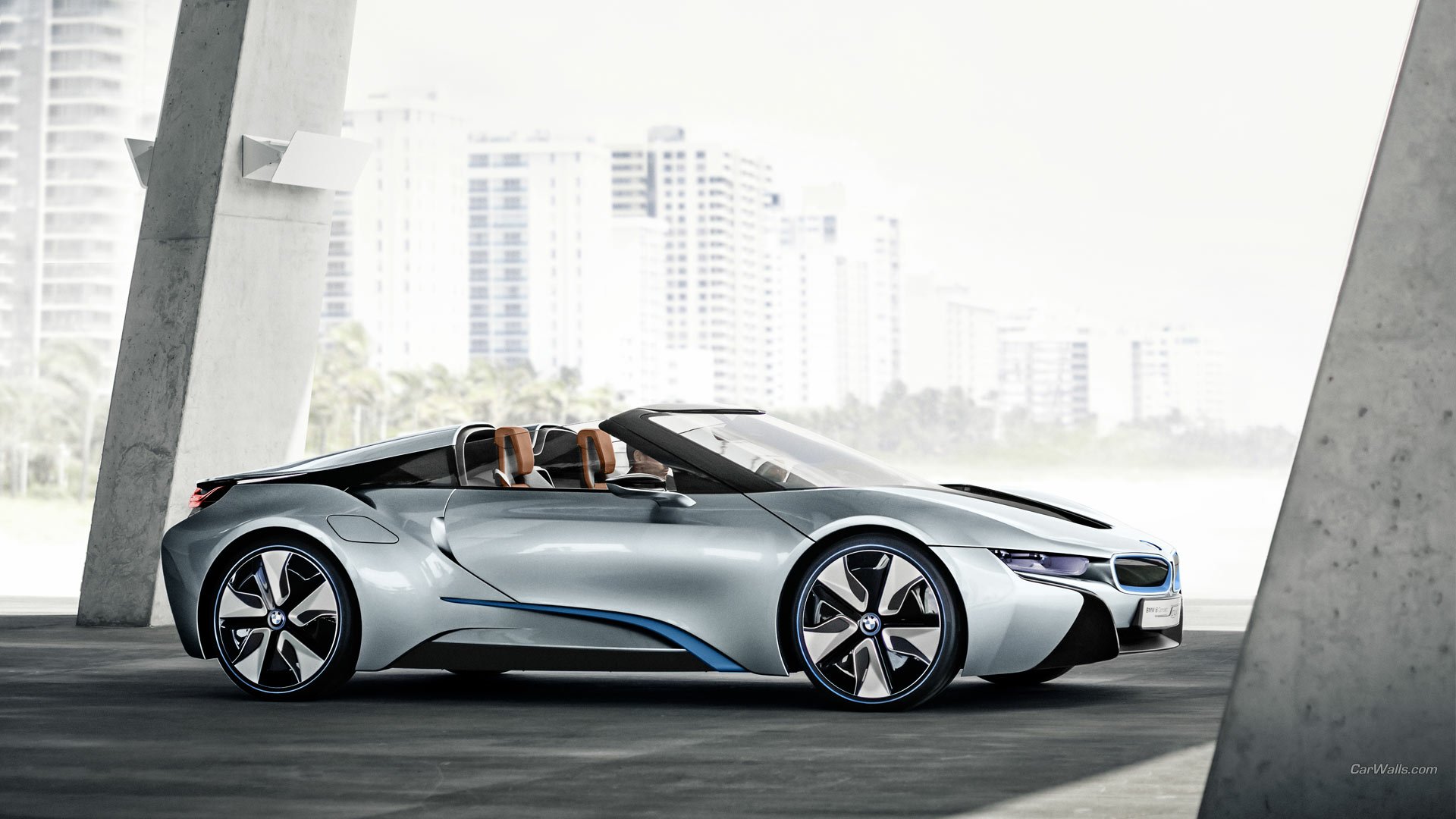 Sleek silver BMW i8 Concept Spyder roadster in profile under a modern overpass, sunlit city skyline in the background — HD PC desktop wallpaper/background.