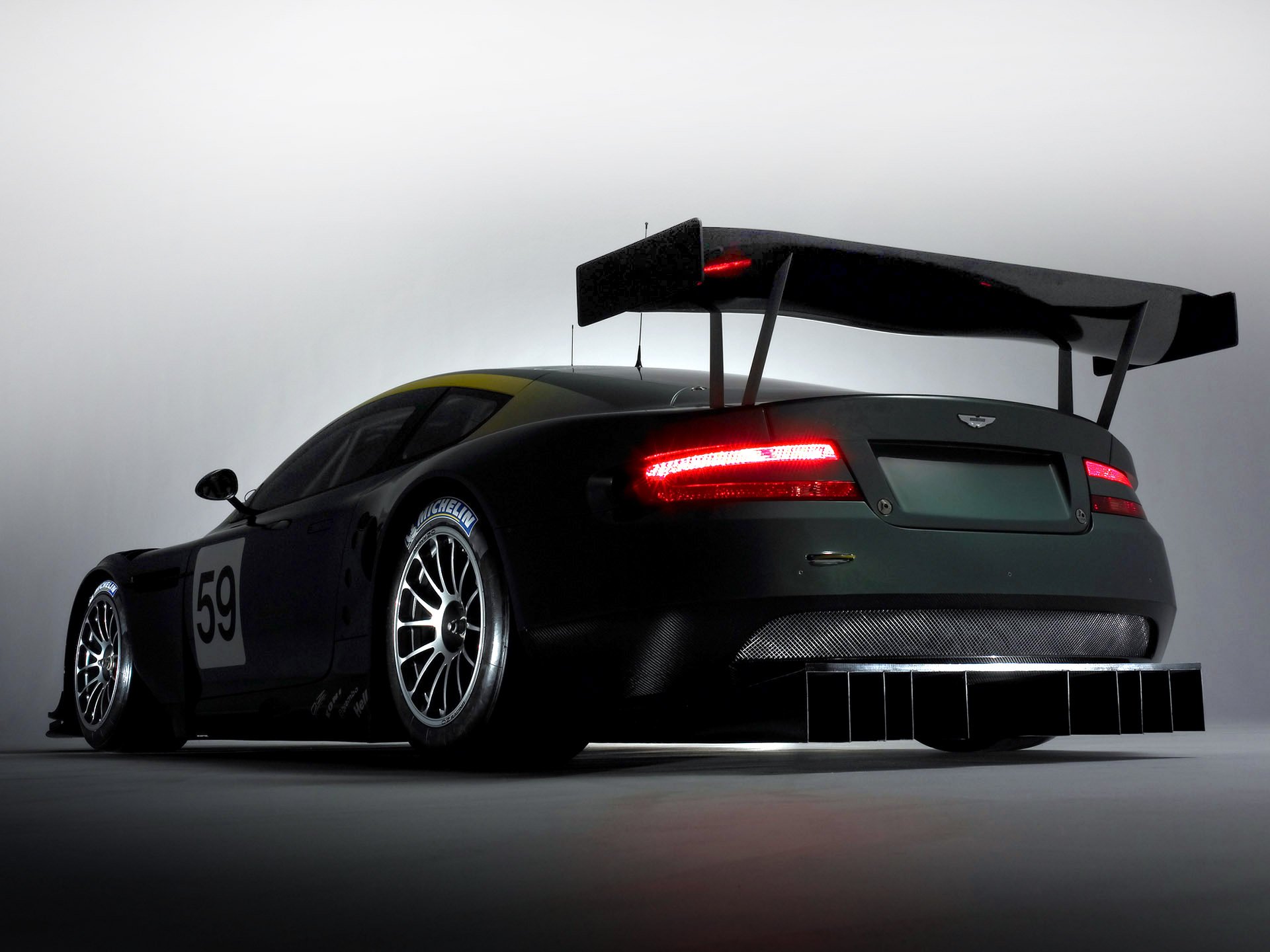 HD PC desktop wallpaper featuring an Aston Martin DBR9 race car in rear three-quarter view, glowing taillights and large rear wing against a minimalist gray background.