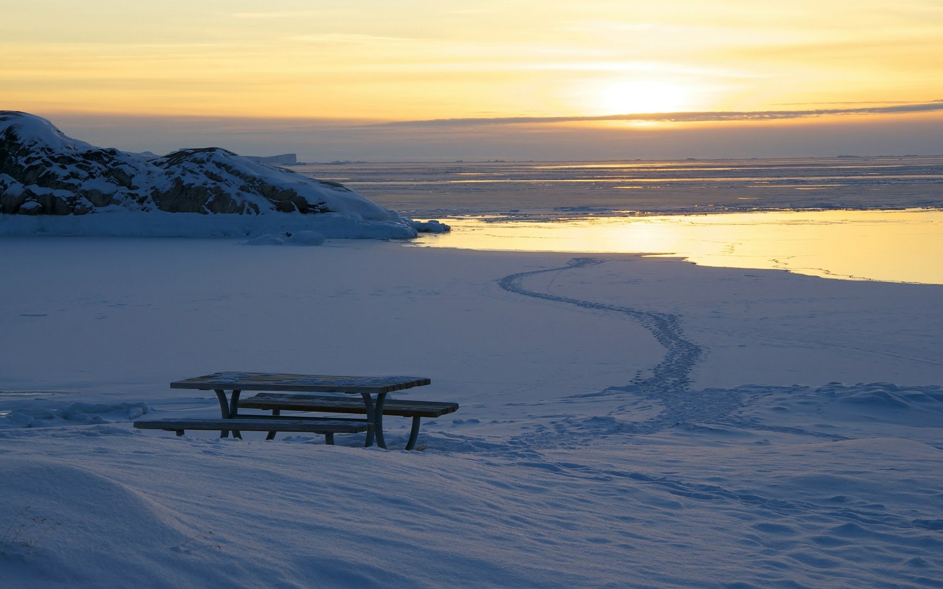 HD PC desktop wallpaper — winter sunset photography of a snow-covered shore with a picnic bench beside an icy sea, golden sun reflecting on calm water.