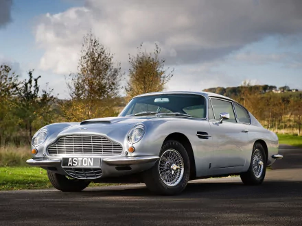 HD PC desktop wallpaper of a silver Aston Martin DB6 vehicle parked on a country road, three-quarter front view under a cloudy sky.