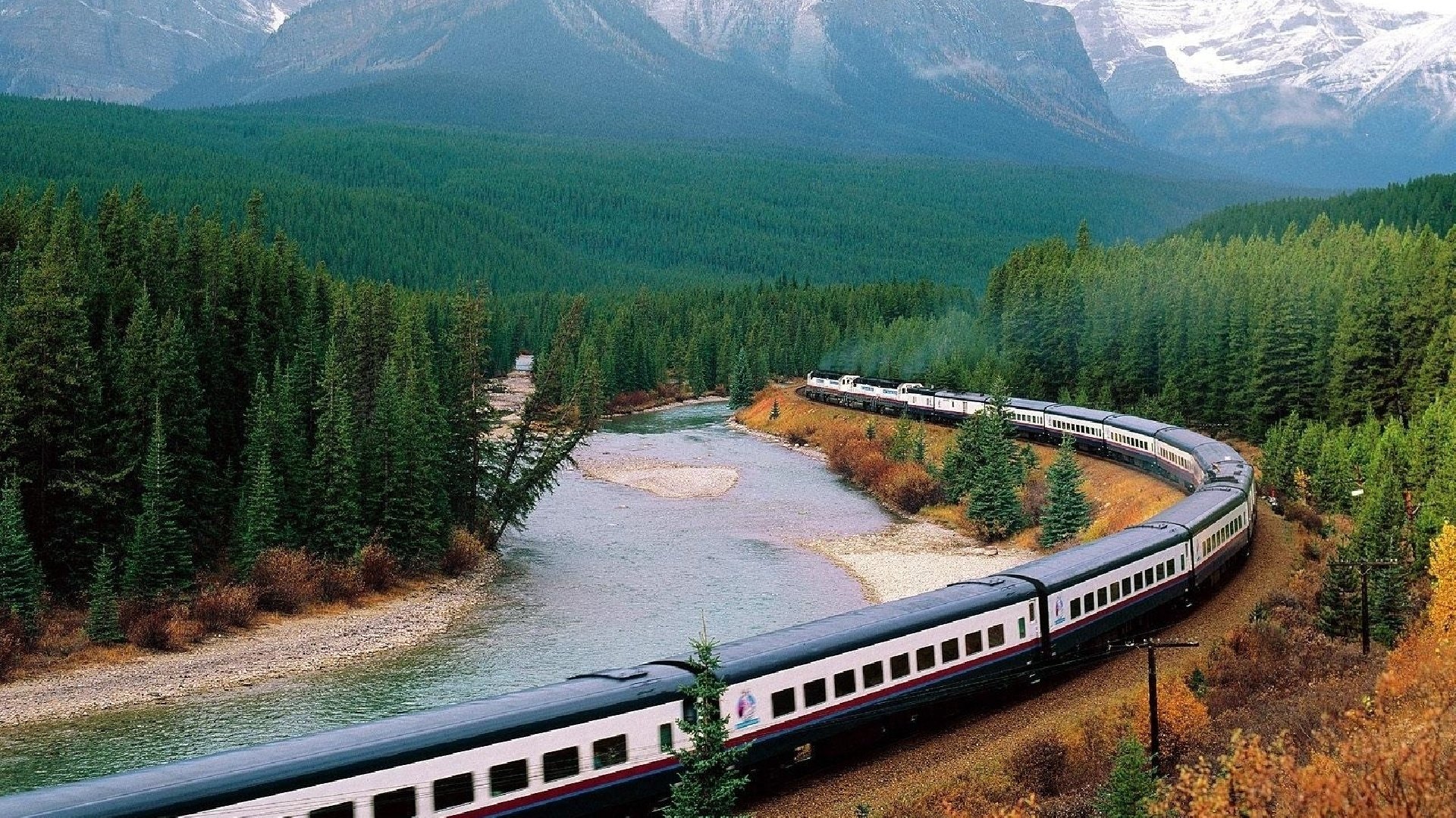HD PC desktop wallpaper showing a train curving along a river through a dense forest with mountains in the background under a cloudy sky.