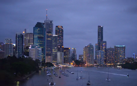 A serene dusk view of Brisbane, Queensland, showcasing the city skyline with shimmering lights reflected on the river, dotted with sailboats and surrounded by modern buildings.