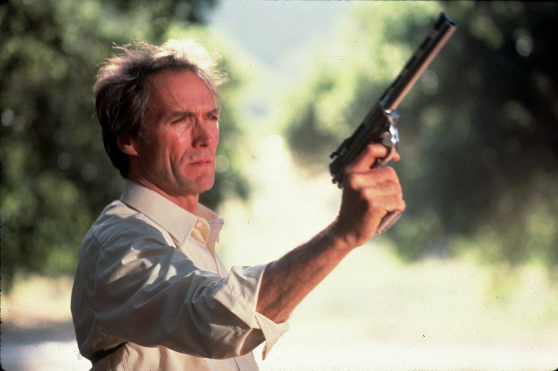 Movie still (Sudden Impact): actor in a light shirt aims a revolver outdoors, sunlit foliage behind him — 2K Quad HD PC desktop wallpaper/background.