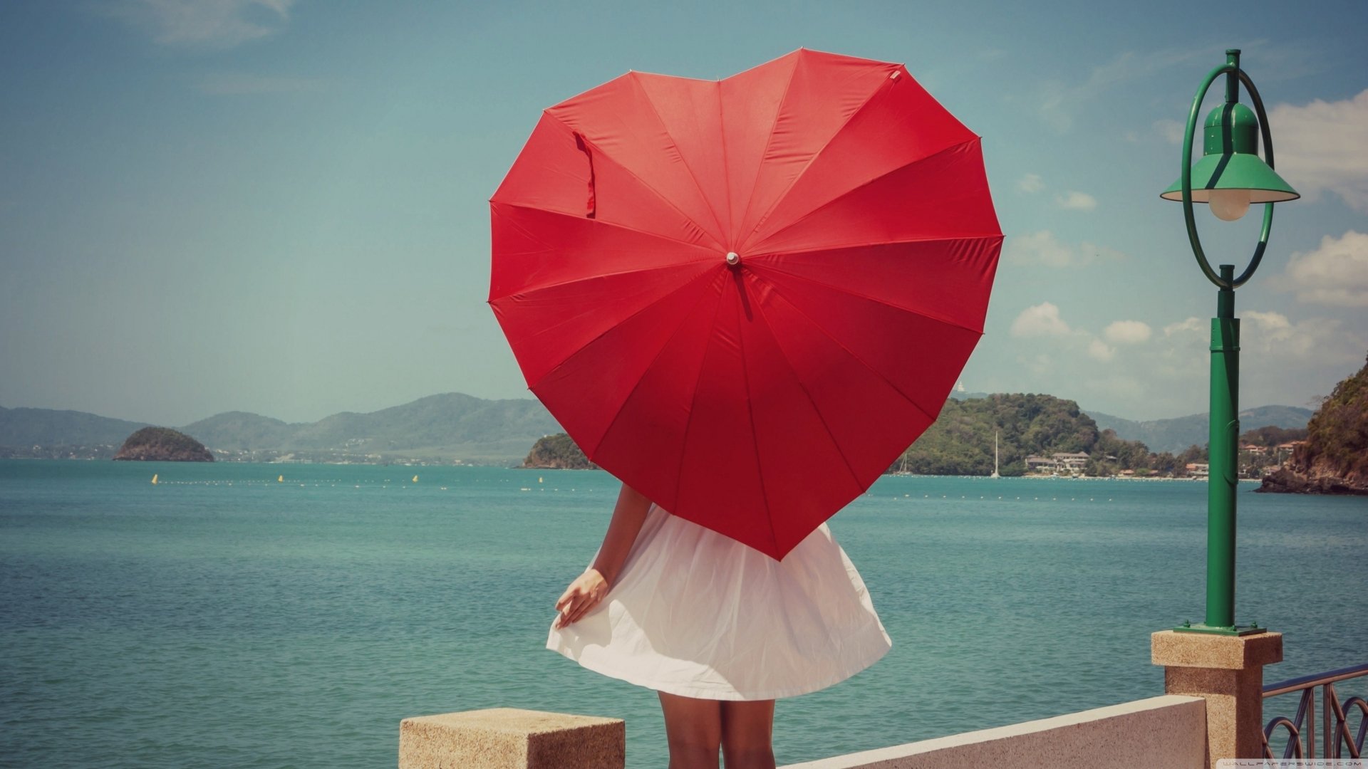 A woman in a white dress stands by the water holding a red heart-shaped umbrella, captured from the rear against a clear sky and distant hills. HD desktop wallpaper.