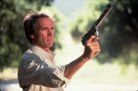 Movie still (Sudden Impact): actor in a light shirt aims a revolver outdoors, sunlit foliage behind him — 2K Quad HD PC desktop wallpaper/background.