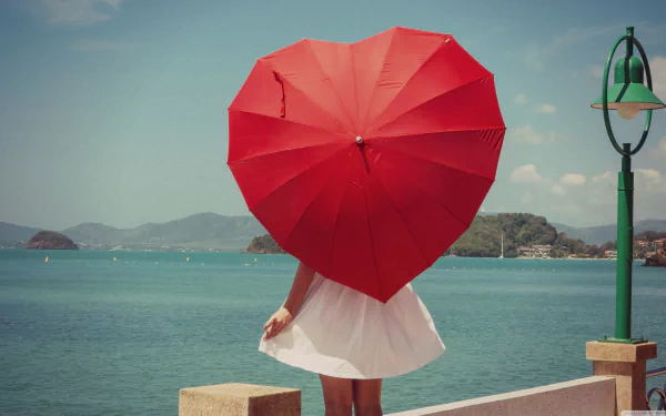 A woman in a white dress stands by the water holding a red heart-shaped umbrella, captured from the rear against a clear sky and distant hills. HD desktop wallpaper.