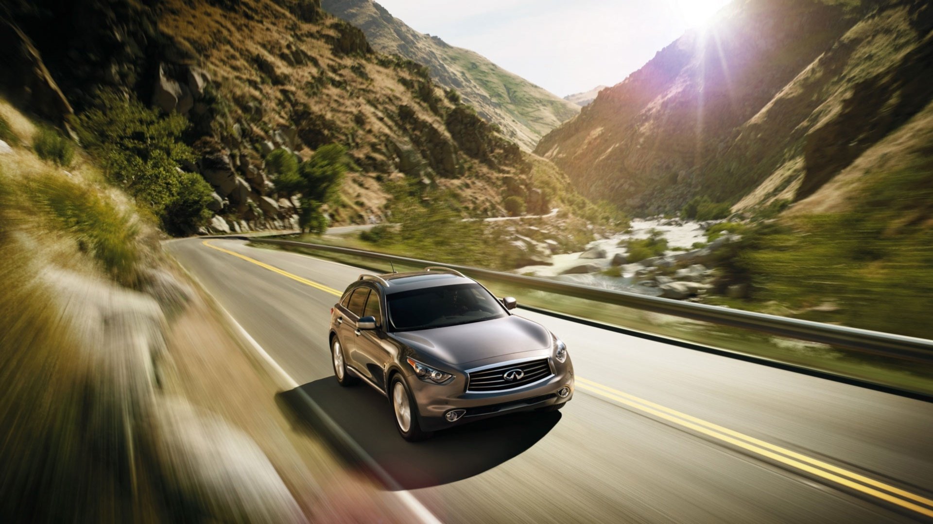 A sleek Infiniti QX70 driving on a winding mountain road, captured in a vibrant HD PC desktop wallpaper with sunlight illuminating the scenic landscape.