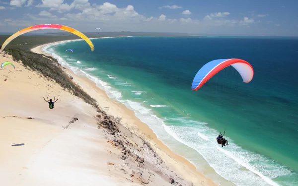 A stunning view of paragliders soaring over a sandy beach and vibrant ocean, capturing the thrill of the sport in a picturesque HD desktop wallpaper.
