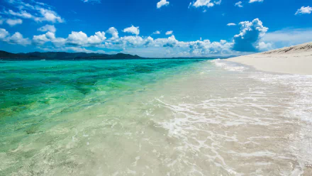 A serene seascape featuring clear turquoise waters gently lapping against a sandy shore under a bright blue sky with fluffy clouds, captured in vibrant 4K Ultra HD for a stunning desktop wallpaper.