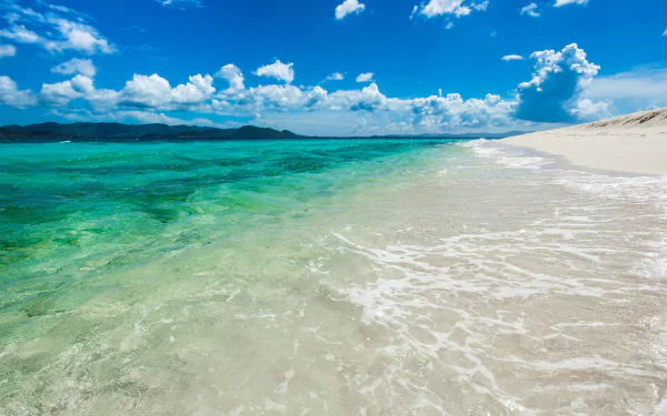 A serene seascape featuring clear turquoise waters gently lapping against a sandy shore under a bright blue sky with fluffy clouds, captured in vibrant 4K Ultra HD for a stunning desktop wallpaper.
