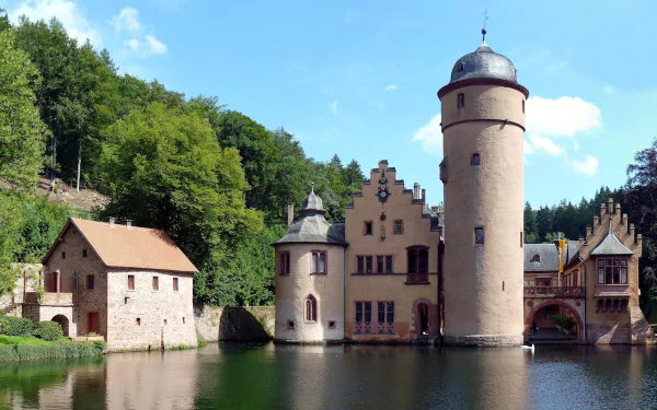 Mespelbrunn Castle in Germany, a historic man-made fortress surrounded by water and lush greenery under a clear blue sky, shown in HD desktop wallpaper quality.
