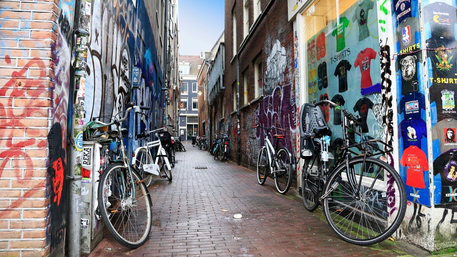 HD PC desktop wallpaper of a man-made Amsterdam alley: brick-paved lane flanked by graffiti-covered walls and parked bicycles between narrow buildings.