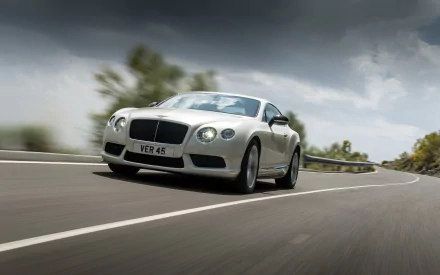 Silver Bentley Continental GT V8 racing along a curving road beneath stormy skies — 2K Quad HD PC desktop wallpaper and background.
